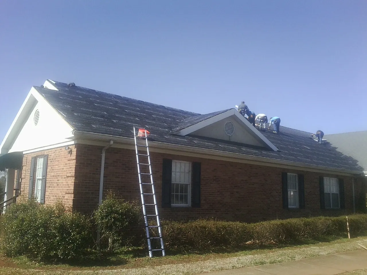 Crew tearing shingles off a brick ranch roof with a ladder set — Emergency Roof Repair work by our crew in Burlingham