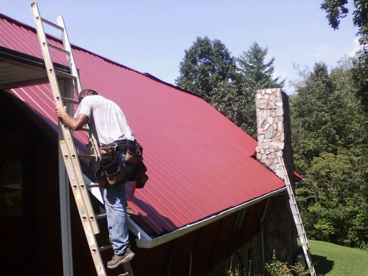 Roofer on a ladder beside a red metal roof and stone chimney — Soffit & Fascia Repair by Roofing Brothers in Burlingham